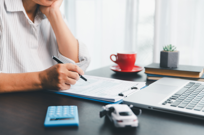 Person sitting down at desk holding a pen signing documents with a calculator and toy car right next to them.