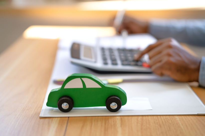 Arms at a desk covered with papers punching numbers into a calculator with a green car at the front of the desk