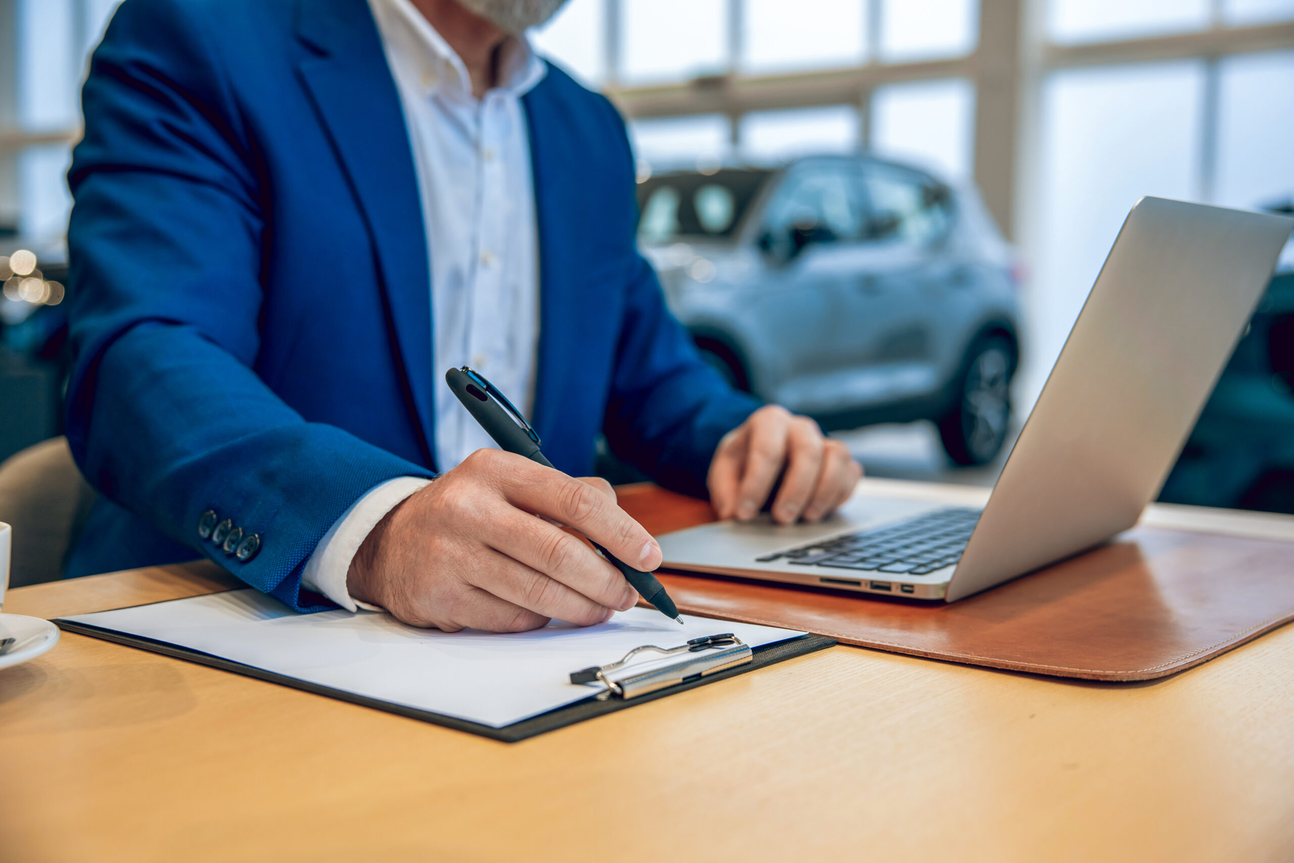 Person in a suit sitting at a desk in a car dealership looking at a laptop with paperwork on the desk.