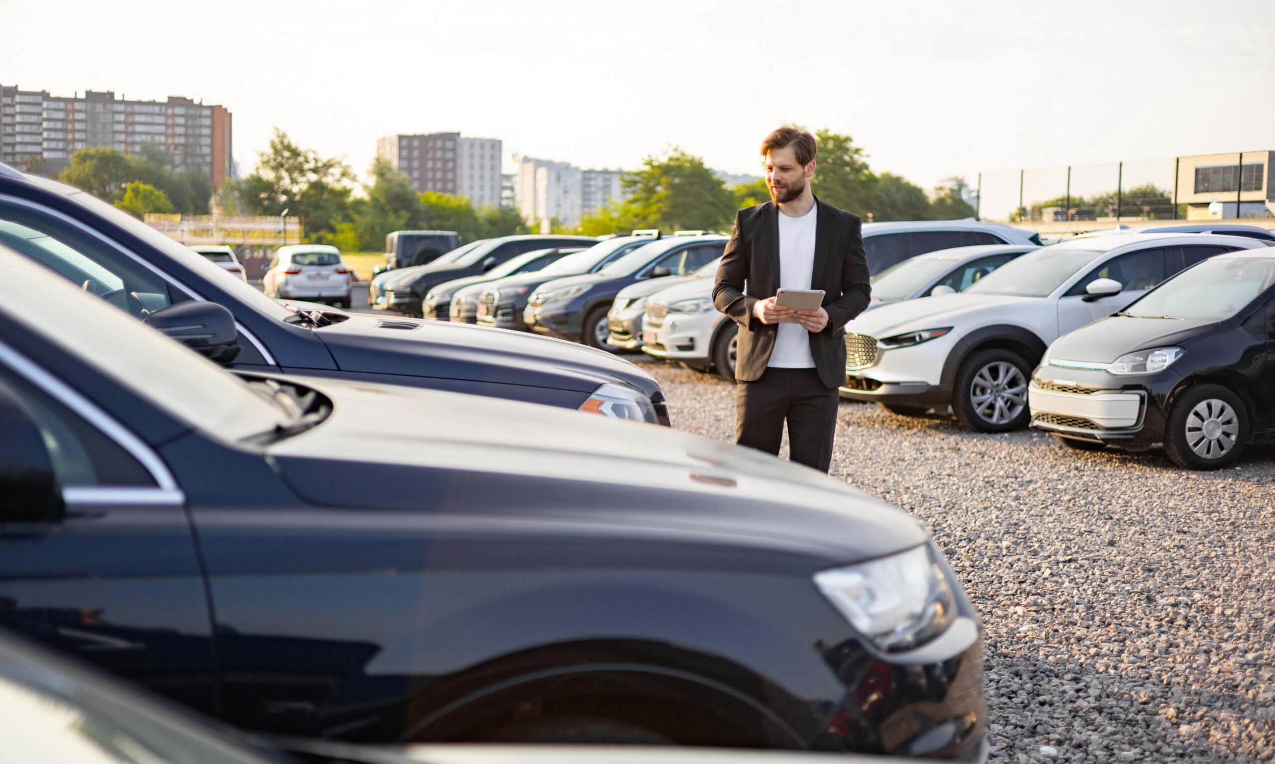 A car dealer in a suit using a tablet while standing in a car dealership, surrounded by rows of new cars on a sunny day.