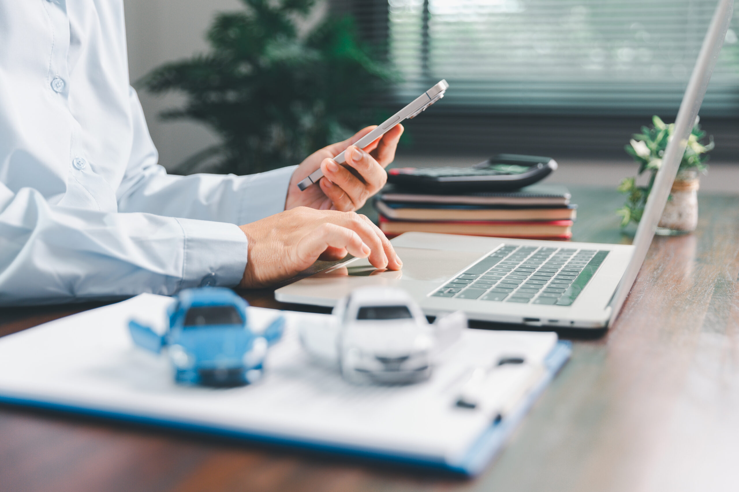 Pair of hands at a desk holding a cell phone and typing on a computer with notepad and toy cars in the front