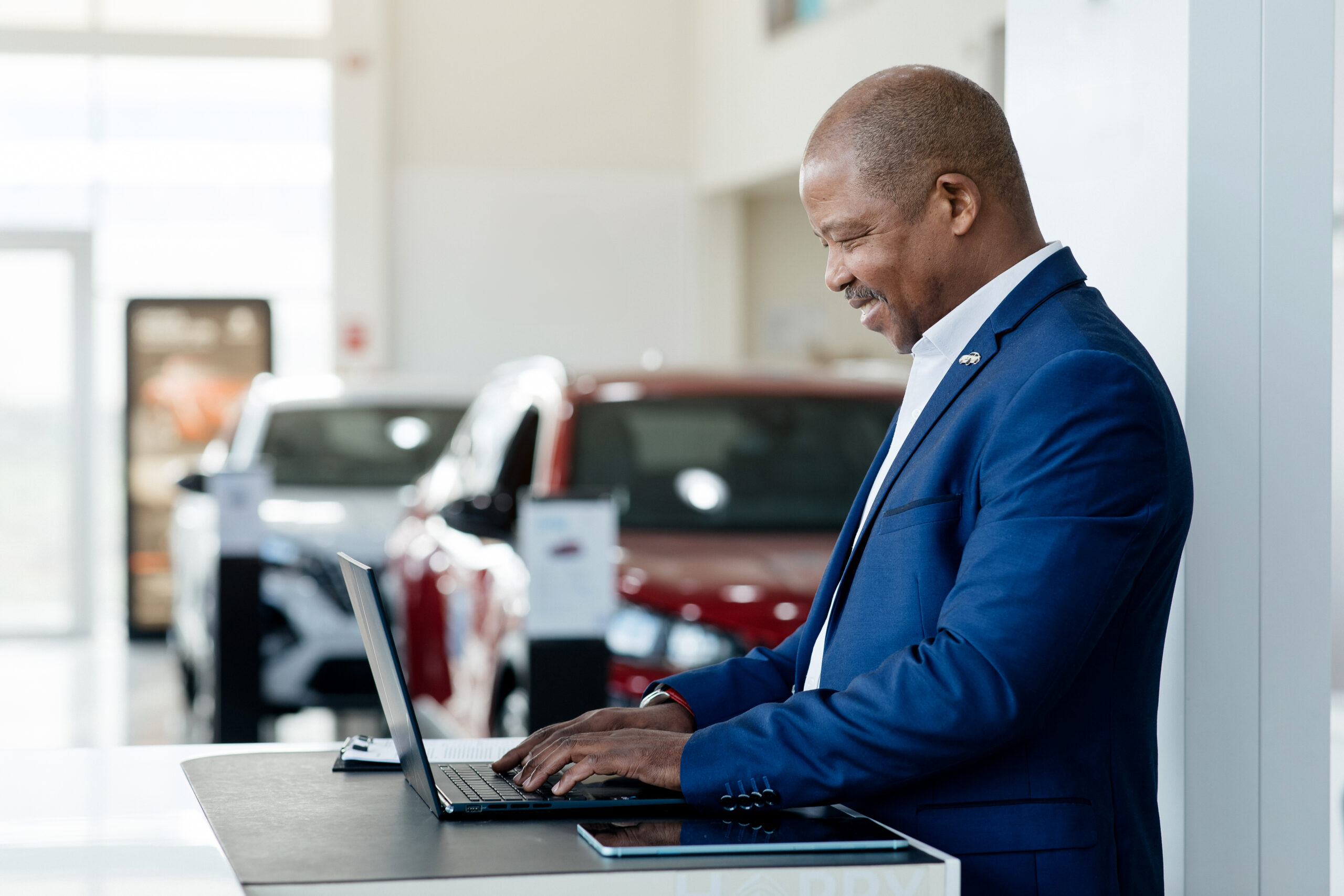Positive car salesman using laptop computer at work desk in auto dealership.