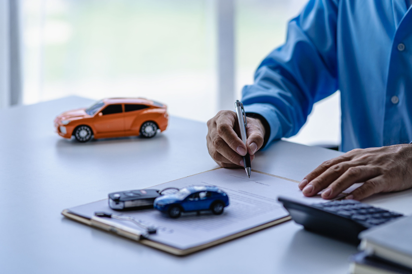 Man writing on a piece of paper. On the desk next time him is a small orange car and a car key.